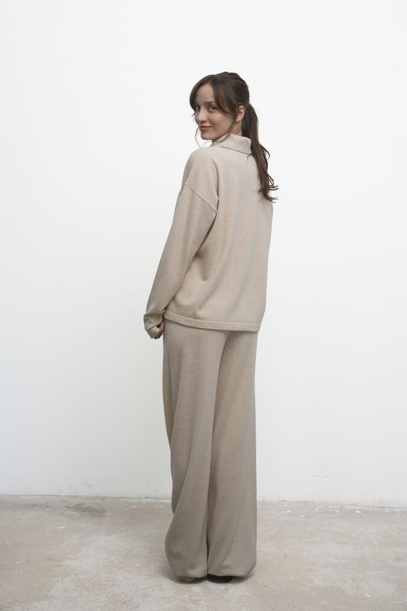 Sweter Lorenza Beige – beżowy sweter damski, tyłem, modelka odwraca głowę do aparatu, beżowe spodnie