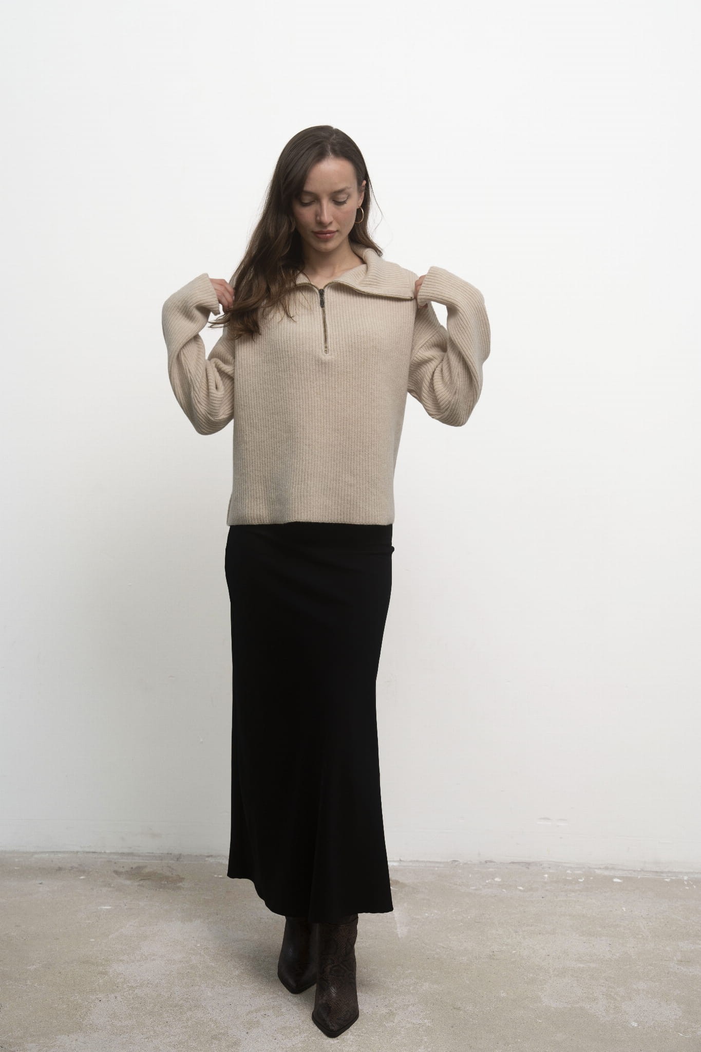 Sweter Luna Beige – beżowy sweter damski, przodem, modelka trzyma się za ramiona, czarna spódnica i kozaki